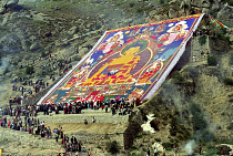 Parade toward the Thangka and onlookers at a silken Thangka Buddhist ceremony for the cycle of life with massive brightly colourd hillside Buddha image