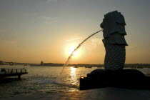 Merlion statue spouting water in silhouette overlooking Singapore River at sunset