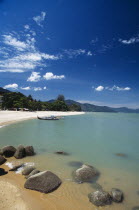 View along shore of quiet sandy beach with moored boat in middle distance and smooth rocks in shallow water in the foreground.