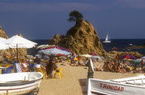 Busy sandy beach with people sitting at tables under umbrellas or sunbathing.  Central rocky outcrop and boats on beach in foreground.