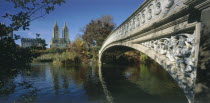 Central Park  Bow Bridge.  View along side of cast iron bridge across lake lined by trees and overlooked by city buildings and the San Remo towers .