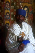 Church of Debre Birhan Selasie.  Portrait of priest in front of painted wall  holding crucifix.
