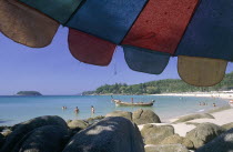 Little Kata Beach seen from beneath a colourful umbrella with rocks  a boat and people wading in the sea