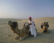 Two Thar Desert camels lying down in the sand  with a man wearing a pink turban leaning against one