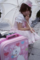 Harajuku District. Portrait of young teenage girl wearing a pink and white dress holding a parasol with a pink suitcase in the foreground
