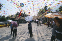 People walking on a path holding umbrellers in Daguan Park with colourful flags overhead.