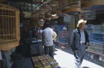 A man wearing a hat walking through a alley way in a Bird and Flower market surrounded by bird cages.