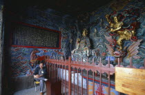 Interior view of the temple beside Dragon Gate in Western Hills with brightly painted walls and golden statuary and a man sat on a chair.