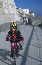 Children, Playing, Young girl on a bicycle dressed in protective cycling helmet and pads riding on a path next to the beach.