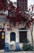 Kardamili. Taverna exterior with blue table and chairs on pavement and red flowering tree growing against the building.