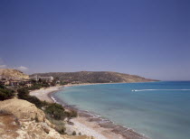 Water skier and speed boat in Pissouri Bay encircled by quiet beach with surrounding rocky coastline overlooked by hotel and other buildings.