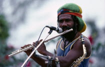 Rastafarian musician singing and playing flute at a concert.