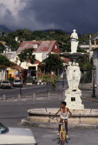 Large white statue in middle of street surrounded by houses and a boy on a bike in the foreground.
