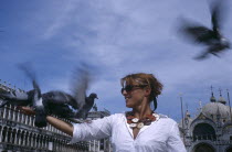 St Marks Square. Woman wearing a white shirt standing with pigeons fluttering in motion blur