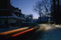 Streaked red car lights in motion blur on road through snowy village at dusk