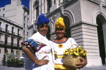 Two smiling women wearing brightly coloured head scarfs standing in the square holding a basket of flowers