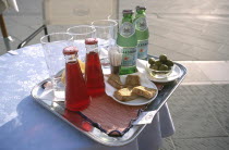 Tray on a small round lace clothed table with glasses and two bottles of Mineral water standing with two bottles of Campari and a dish of Olives