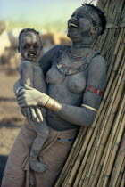 Agar Dinka woman decorated with dust  holding young son in her arms and laughing. Woman wearing marriage beads around her neck and white ivory bracelet on her arm.