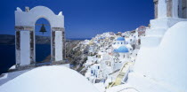 Oia.  White painted hill town with bell in foreground hanging from arch part framing view over sea to coastline.
