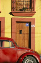 Partly seen red volkswagon beetle outside yellow building with wooden door and window shutters in orange painted frames.