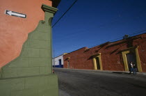 Corner of green and orange painted building with white arrow on black background pointing left.  Long orange building with yellow door frames across road behind.