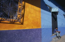Part view of building painted in rectangles of ochre  blue and purple with partly open purple metal window screen  casting shadow.  Boy sitting in doorway.