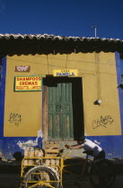 Man pushing bicycle handcart stacked with crates of fruit past yellow and blue painted building facade with advertisment for shampoo beside green door.