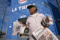 Male newspaper vendor  threequarter view seen from below looking up.  Advertising for Nestle and purified water on blue painted wall of building behind.