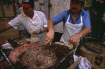 Men cooking meat for tacos and enchiladas on circular hot plate at roadside stall.