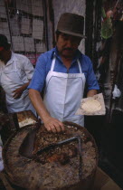 Man making tacos and enchiladas  chopping meat with a cleaver on a circular block.