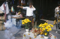 Chinese New Year.  Man at roadside stall holding his ears as he sets off chain of fire crackers.  Table with flowers  incense and food offerings beside him.
