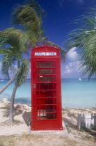 Red telephone box on sand surrounded by palm trees with sea behind.