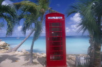 Red telephone box on sand surrounded by palm trees with sea and distant boat behind.