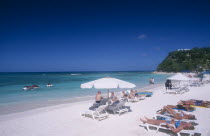 Beach overlooking semicircular bay with sunbathers on white sun loungers.  Two jet skis in the water and tree covered headland behind.