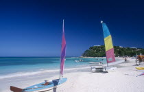 Semi circular bay beside quiet beach  boats with colourful sails pulled up onto the sand and motor boats and jet skis moored in the water.  Tree covered headland behind.