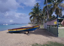 Empty sandy beach fringed by palm trees with painted wooden boat pulled up out of the water beside empty hut advertising Carib beer.
