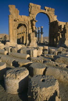 Monumental arch. High central arch flanked by lower arch on each side with colonnaded street partly seen behind and masonry ruins in the foreground.