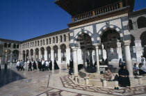 The Umayyad Mosque.  The ablutions fountain with pilgrims sitting around the hexagonal base and crossing the marble courtyard floor at its left.