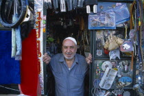 Male shopkeeper at the entrance to his shop with display of mixed goods hanging from the walls and ceiling.