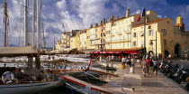 View along busy waterfront  with boats moored at the harbourside  open-fronted restaurants and the local tourist office.  Var.