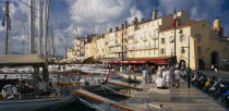 View along busy waterfront  with boats moored at the harbourside  open-fronted restaurants and the local tourist office.  Var region.
