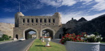 Muscat Gate Museum   Crenellated gatehouse with archways over roads divided by central strip of grass with  white tubs of red and white flowers. Learning Lessons Middle East Omani Teaching