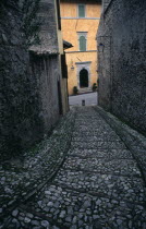 A narrow cobbled street leading down towards a yellow painted building partly seen between grey stone walls.European Gray Italia Italian Southern Europe