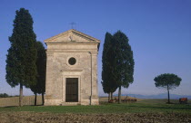 Small stone chapel  facade with cypress trees growing beside it in an arable landscape.