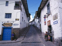 View up a narrow street between white painted houses.   Woman and child in peasant dress standing opposite a woman in modern dress at the bottom.