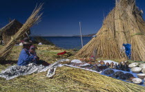 Woman and child seated outside a reed house on a floating island  decorative wares displayed beside them.