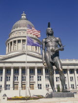 Utah State Capitol.  Detail of dome and colonnaded facade.  Flags flying from flagpole and Indian statue in the foreground.