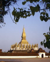 Pha That Luang Sacred Stupa with gold spires and white walls part framed by tree branches.