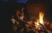 Zulu man making a spear over an open fire at night outside thatched huts