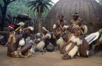 Tribesmen sitting on the ground outside a hut whilst the chief sits on a chair because of his seniority